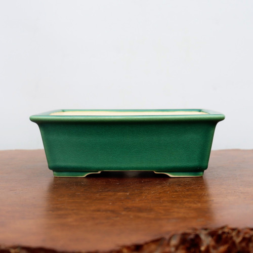A detailed, eye-level shot of a rectangular, forest green glazed ceramic bonsai pot resting on a polished, warm-toned wooden surface. The pot features a thick, rounded top rim with sides that taper slightly inward toward the base. Its vibrant green glaze has a high-gloss finish and a visible fine crackle or "crazing" pattern across the entire exterior. The pot is supported by short, integrated feet at the corners, creating a small arched space underneath for drainage and airflow. The minimalist background consists of a plain, off-white wall, which emphasizes the deep green color and craftsmanship of the container.