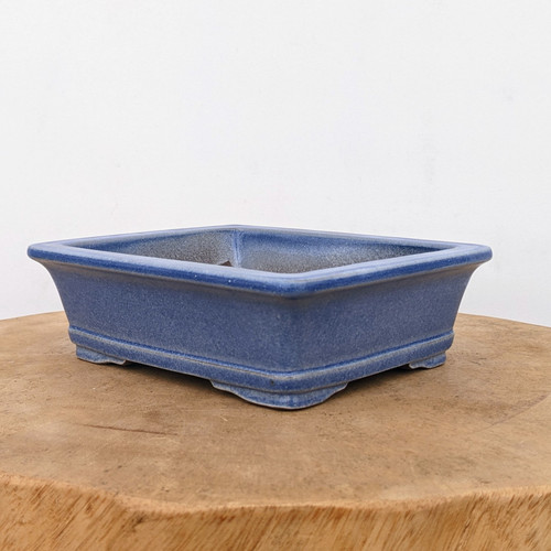 A high-resolution product photograph shows a seven-inch rectangular glazed bonsai pot in a deep, vibrant cobalt blue. The ceramic pot features elegantly flared sides that sweep upward to a prominent, rounded lip at the rim. Its surface has a subtle, mottled texture within the glossy blue glaze, providing a rich, artisanal feel. The pot is supported by four small, decorative cloud-style feet at the corners, elevating it slightly above the surface. The interior of the pot appears unglazed with a grainy, earth-toned texture, suitable for healthy root development. The pot is presented on a thick, circular slab of natural-grained light brown wood, set against a clean, minimalist white background that makes the bold blue color and elegant silhouette of the bonsai container stand out.