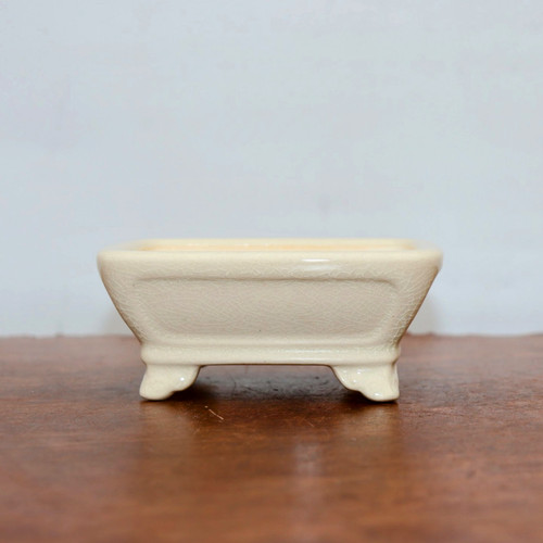A studio photograph shows a side view of a 5-inch Cream Glazed Yixing Bonsai Pot (model YX2043). This rectangular ceramic pot features a smooth, off-white cream finish with a fine crackle glaze (craquelure) adding texture to its surface. The design includes outward-sloping sides with a recessed rectangular panel detail on the front and back. The pot is supported by four decorative, traditional-style feet at each corner, creating a small space between the container and the surface. It is centered on a polished, dark reddish-brown wooden tabletop against a neutral, light-colored background, highlighting the elegant and clean aesthetic of this miniature bonsai container.