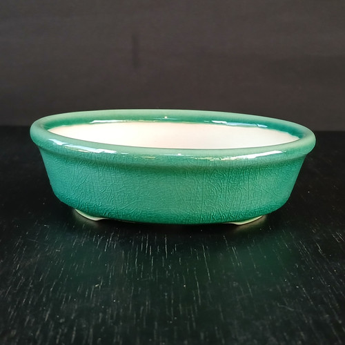 A close-up photograph of a shallow, oval-shaped glazed bonsai pot. The exterior is finished with a vibrant green crackle glaze, featuring a delicate network of crazing lines that add a vintage texture. The pot's interior is a contrasting smooth, off-white color. It has a classic rolled rim and is supported by small, discreet feet at the base. The pot is positioned on a dark, textured black surface against a plain, dark gray background, which makes the bright green glaze stand out as the central focus.