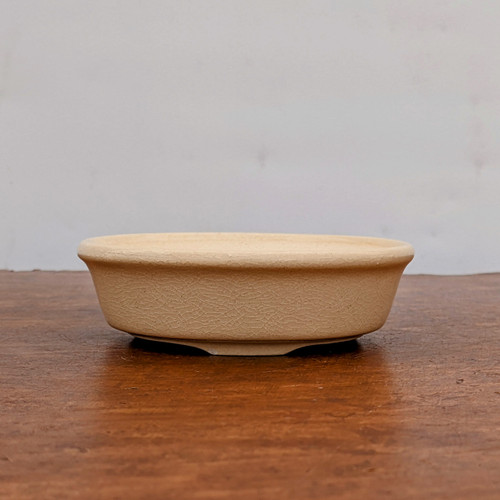 A detailed, eye-level side view of a shallow, cream-colored ceramic bonsai pot, designated as model "Cream 2005." The pot features a rounded, oval shape with a slightly flared rim and a small, integrated footed base. Its most distinctive feature is a fine, intricate crackle glaze (crazing) that covers the entire exterior, giving the light beige surface a textured, antique appearance. The pot is centered on a dark, weathered wooden tabletop that shows natural grain and some distressing. The background is a plain, neutral light gray wall, creating a clean and minimalist composition that emphasizes the pot's shape and finish. The soft, even lighting highlights the delicate details of the glaze and the sturdy form of the ceramic container.