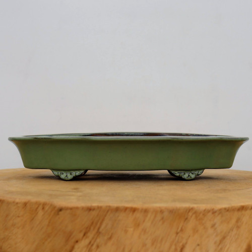 An eye-level studio photograph features an eleven-inch rectangular bonsai pot with a muted green glaze, resting on a rustic wooden slab. The shallow ceramic planter has a wide, slightly flared upper rim and is finished in an earthy olive-green tone with a smooth, matte-like texture. Two visible ornate feet support the pot, each intricately carved with traditional decorative scrollwork. The pot is centered on a thick cross-section of a natural wood log that showcases a warm, vertical grain pattern. The background is a clean, minimalist off-white, focusing all attention on the craftsmanship and low-profile design of the bonsai container.