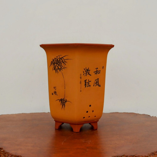 An eight-inch, tall, square unglazed bonsai planter with a tapered body and elegant flared rim. The pot features a warm, ochre-colored matte finish on its main body, contrasted by a dark reddish-brown clay on the rim and four short, blocky feet. The front of the planter is adorned with hand-etched artwork: a delicate, slender bamboo plant on the left and bold calligraphic characters on the right. A set of small drainage holes is arranged in a decorative triangular pattern near the base. The pot is shown resting on a polished, dark burl wood surface against a neutral white background, highlighting its traditional and artistic design.