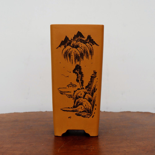 A tall, five-inch rectangular unglazed bonsai pot in a warm ochre-orange hue, featuring a detailed black-etched landscape. The etching showcases a traditional East Asian scene with jagged mountain peaks, a large weeping willow tree, and a small pavilion nestled among rocky outcrops. The slender planter is shown standing on a dark wooden table against a plain white wall, highlighting its elegant shape and artisanal hand-drawn artwork. This type of etched ceramic pot is ideal for accentuating the natural beauty of a small bonsai tree.