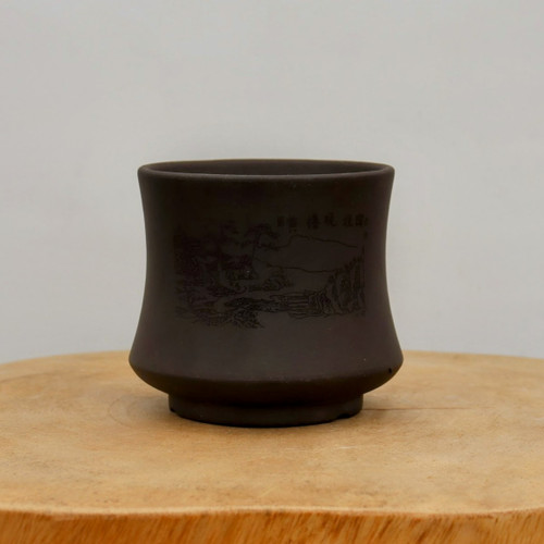 An elegant, dark brown unglazed bonsai pot with a subtle hourglass shape sits on a light-colored wooden surface against a neutral, off-white background. The small, cylindrical pot is made of matte-finished clay and features an intricate landscape scene etched into its front. This traditional East Asian-style illustration includes mountains, pine trees, and water, with several calligraphic characters engraved next to the scene. The pot has a slightly flared rim and a small, circular footed base. The earthy tone and delicate etching of the unglazed ceramic provide a classic and sophisticated home for a miniature bonsai tree.