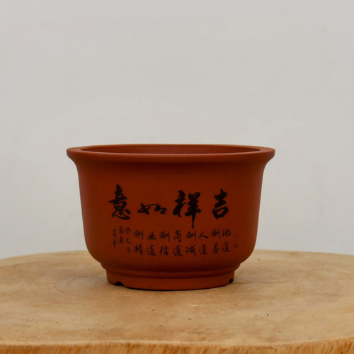 A studio photograph captures a deep, round, unglazed bonsai pot crafted from reddish-brown terracotta. The pot has a traditional silhouette, tapering slightly from a flared rim down to a sturdy, raised base. 

The front of the pot is decorated with elegant black calligraphy. Four large, expressive characters are centered, reading "吉祥如意" (Jí xiáng rú yì), a traditional Chinese blessing for good luck and prosperity. Beneath these main characters are several columns of smaller, finely etched script, which traditionally detail philosophies of ethics, business, and nature. 

The pot is presented on a light-grained, circular wooden platform, which contrasts with its warm, earthy clay tones. The background is a minimalist, solid off-white wall, ensuring that the focus remains entirely on the pot's texture and artistic detailing. This etched bonsai pot offers a classic and culturally rich vessel for a variety of bonsai tree styles.