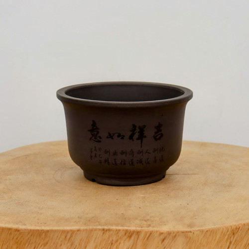 A dark brown, unglazed ceramic bonsai pot sits centered on a thick, light-colored natural wood slab. The pot features a classic rounded, bowl-like shape with a subtle flared rim and a sturdy, slightly raised footed base. Its smooth, matte surface is adorned with traditional Chinese calligraphy etched into the clay and filled with black ink. 

The four prominent characters in the center are written in a fluid, artistic script, translating to "Jíxiáng rúyì," a classic blessing for good fortune and may all things go according to one's wishes. Below this main inscription are smaller vertical columns of more delicate characters, which list various philosophical virtues such as sincerity and professional excellence. The pot’s earthy, dark chocolate tone provides a stark contrast to the pale, grainy wood of the stand and the clean, neutral light-gray background. This traditional piece of pottery exemplifies the understated elegance and cultural artistry often sought in bonsai cultivation.
