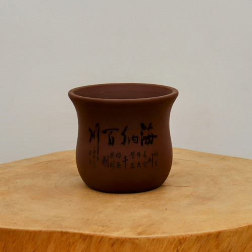 An unglazed bonsai pot in a rich reddish-brown clay, sitting on a rustic, light-colored natural wood pedestal. The pot features a rounded, tapered shape with a subtle flare at the top rim. Etched into the side of the pot is traditional Chinese calligraphy in black ink, including four large, prominent characters and a smaller inscription beneath them. The scene is set against a clean, neutral off-white background, highlighting the earthy tones of the pottery and the organic texture of the wood grain on which it rests.