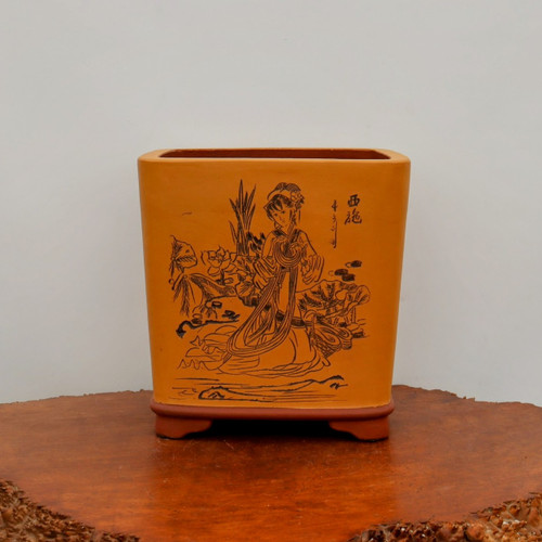 A rectangular, unglazed bonsai pot crafted from warm mustard-yellow clay, resting on a reddish-brown ceramic base with four small feet. The front face of the pot features a detailed black ink-style etching of a woman in traditional Chinese attire, shown kneeling or seated in a serene landscape with reeds and lotus-like plants. To the right of the figure, several Chinese calligraphy characters are etched into the surface. The pot has a slightly tapered shape with rounded corners and is displayed on a polished, natural-edge wooden surface against a plain, light-colored wall, emphasizing its artisanal and elegant design.
