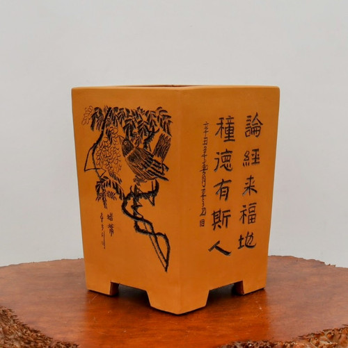 A small, square-shaped unglazed bonsai pot crafted from warm ochre-colored clay sits atop a rustic, polished dark-wood slab. The pot features a classic, upright design with slightly tapered sides and four short, blocky rectangular feet. 

The front face of the pot is decorated with intricate black-etched artwork. On the left side, a detailed illustration depicts a small bird perched on a gnarled, twisted branch surrounded by delicate leaves or blossoms. To the right of the bird, two prominent vertical columns of Chinese calligraphy are carved into the clay, accompanied by smaller inscriptions and a seal near the bird illustration. The matte finish of the unglazed ceramic provides an earthy, traditional aesthetic, highlighting the fine lines of the hand-etched decorations. The entire composition is set against a clean, off-white background, emphasizing the artistic details of this functional bonsai accessory.