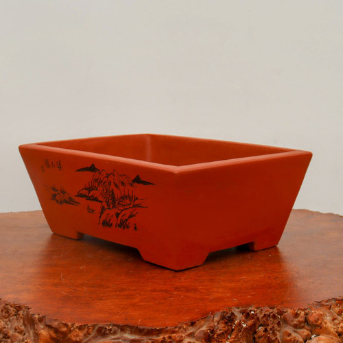 A rectangular, unglazed terracotta-colored bonsai pot sits centered on a polished wood burl base. The pot features a classic design with sloping sides that flare outwards towards the top and rests on four small, rectangular integrated feet. One of the long sides is decorated with a detailed black etched landscape scene in a traditional Chinese style. This illustration depicts jagged mountain peaks, scattered pine trees, a small hut, and calm water. In the top-left corner of the landscape, there are three small, vertically aligned Chinese characters. The pot is showcased on a lustrous, warm-toned wooden slab with a natural, irregular live edge, set against a clean, off-white background.