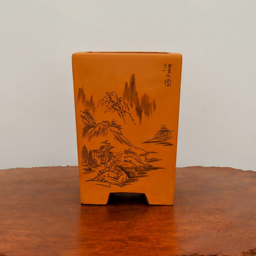 A tall, rectangular unglazed bonsai pot with a warm, ochre-orange terracotta color, featuring a detailed hand-etched landscape design in dark ink. The pot stands on a polished wooden surface with a rich grain, set against a neutral, light gray background. The intricate etching depicts a traditional Chinese-style landscape scene with towering, jagged mountains in the background, rolling hills in the mid-ground, and a variety of stylized evergreen and deciduous trees in the foreground. In the upper right corner of the pot's front face, vertical Chinese calligraphy is delicately etched next to a small circular seal or stamp. The pot has a sturdy, slightly tapered form supported by four short, blocky feet at the corners. The overall aesthetic is rustic and artisanal, highlighting the fine craftsmanship of the landscape etching.