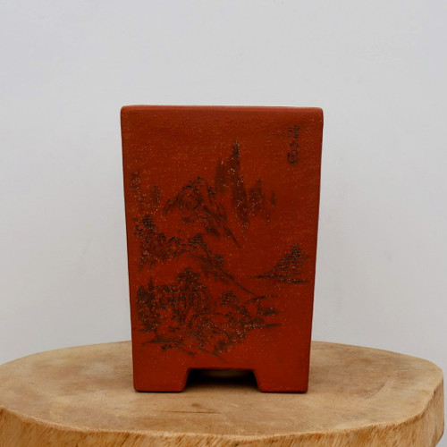A tall, rectangular unglazed bonsai pot made of reddish-brown clay sits centered on a rustic, natural-cut wooden slab. The pot features an intricate, dark-etched landscape scene on its front face, depicting rugged mountain peaks and traditional pine trees in a style reminiscent of classical East Asian ink wash paintings. In the top right corner of the etched face, small vertical calligraphic characters are visible, likely representing an artist's signature or seal. The ceramic surface has a fine, sandy texture, and the pot is supported by four small, integrated feet carved into its base. The entire arrangement is set against a clean, neutral background, emphasizing the earthy tones and artistic craftsmanship of the cascade-style bonsai vessel.