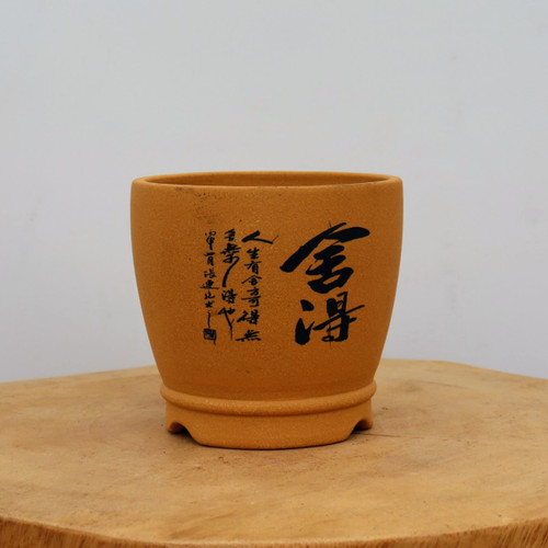 A front-facing view of a round, unglazed ceramic bonsai pot in a warm, light orange-brown earthy tone. The pot features a slightly tapered, bowl-like shape with a raised, multi-lobed base designed for stability and drainage. The front of the pot is decorated with elegant black etched calligraphy. On the right, two large and bold Chinese characters, "舍得" (Shě Dé), are prominently displayed. To the left of these are several vertical columns of smaller, flowing script, with a tiny square red seal mark at the bottom of the text. The pot sits on a natural, light-colored wooden surface with visible wood grain, set against a plain, off-white background with soft, even lighting.