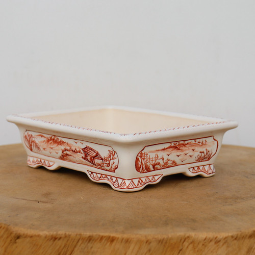 A shallow, rectangular Yixing-style bonsai pot sits atop a thick, natural wood slice against a plain white background. The pot is crafted from a smooth, cream-colored ceramic and features intricate hand-painted artwork in a warm, reddish-brown russet tone. 

On the long side of the pot, a framed landscape scene depicts a traditional countryside setting with a small cottage nestled among rolling hills, stylized trees, and birds soaring in the sky. The shorter side features a similar scenic panel with a dense forest and a distant horizon. 

The pot's design includes a wide, flat rim decorated with a delicate cross-hatched border. At the base, the pot is supported by scalloped, decorative feet, which are further embellished with a geometric, triangular pattern that complements the traditional aesthetic. The overall look is one of refined elegance, combining the functionality of a bonsai container with the artistry of classic landscape painting.