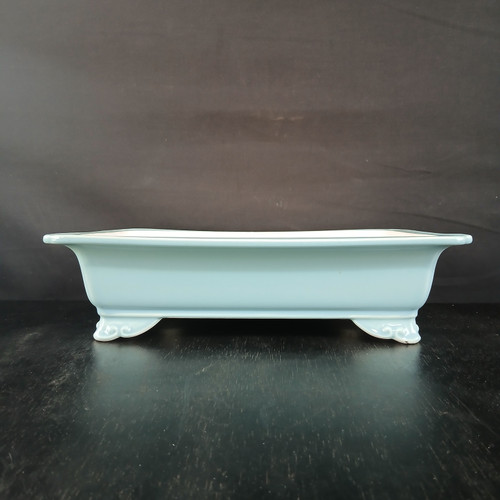 An eye-level, side-profile image shows a rectangular, light blue glazed Yixing bonsai pot. This shallow ceramic planter features a wide, outward-flaring rim and sits on four short, ornate feet with delicate scrollwork carvings. The smooth, glossy glaze is a soft, uniform pale sky blue. The pot is centered horizontally on a dark, textured wooden surface that reflects its bottom edge. Behind the pot is a solid, matte black fabric backdrop, creating a high-contrast setting that emphasizes the pot's elegant form and serene color. This type of high-quality ceramic container is designed for artistic bonsai tree displays.
