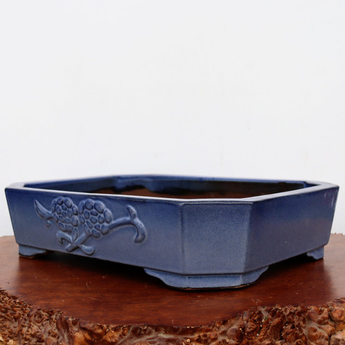 A low-profile, rectangular Yixing bonsai pot with a rich blue glaze, featuring elegantly notched corners and a shallow depth. The ceramic planter is finished in a vibrant indigo or sapphire blue with subtle tonal variations that give the surface a handcrafted, textured appearance. On the front-facing panel, there is a raised, embossed floral or pine needle motif. The pot sits on four low, integrated feet for stability and drainage. Its interior is a dark, unglazed earthy brown, characteristic of traditional Yixing clay. The planter is displayed on a polished, warm-toned wooden slab with a natural, textured burl edge, set against a clean, off-white background.