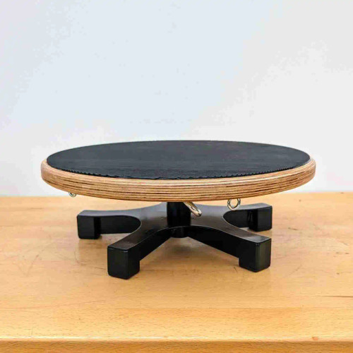 A high-angle, detailed view of a professional-grade bonsai turntable workstand, resting on a light-colored wooden surface against a neutral white background. The stand features a thick, circular platform made from high-quality layered plywood, finished with a textured, matte black non-slip surface to provide a secure grip for bonsai pots during pruning, wiring, and styling. Beneath the outer edge of the rotating wooden platform, several small metal eyelet hooks are visible, designed for anchoring training wires to steady a tree while working. The platform is mounted on a sturdy, four-legged black cross-shaped base made of heavy-duty material to ensure stability as the turntable is rotated. This essential bonsai tool is designed for precision and ease of use, allowing artists to view and work on their trees from every angle.