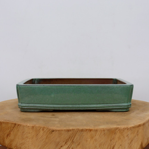 A high-resolution, eye-level photograph shows a ten-inch rectangular Yixing bonsai planter. The pot is finished in a serene, muted seafoam green glaze with subtle darker speckling and earthy brown highlights that accentuate its classic shape and edges. It features a shallow profile with slightly rounded corners and a tiered base defined by two elegant horizontal ridges running around its perimeter. The interior of the pot reveals the unglazed, characteristic reddish-brown clay typical of Yixing pottery. The planter sits prominently on a thick, rustic, natural-edged wooden log slab, all set against a minimalist, clean, light-gray background, emphasizing its design as a sophisticated container for a bonsai tree.