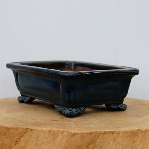 A professional product photograph of a nine-inch dark blue glazed ceramic bonsai pot, shown from a slightly elevated side angle. The rectangular pot is finished in a rich, glossy deep navy blue glaze that reveals subtle texture and tonal variations under soft lighting. Its design features a wide, flared rim and elegant recessed panels on the long sides, adding depth to its form. The pot is elevated by four ornate feet, each intricately carved with a decorative scrollwork pattern. The interior of the pot appears unglazed, showing the natural earthy brown of the clay. It sits centrally on a light-colored, circular wooden block, which is placed against a plain, neutral off-white background to highlight the pot's craftsmanship and classic aesthetic.