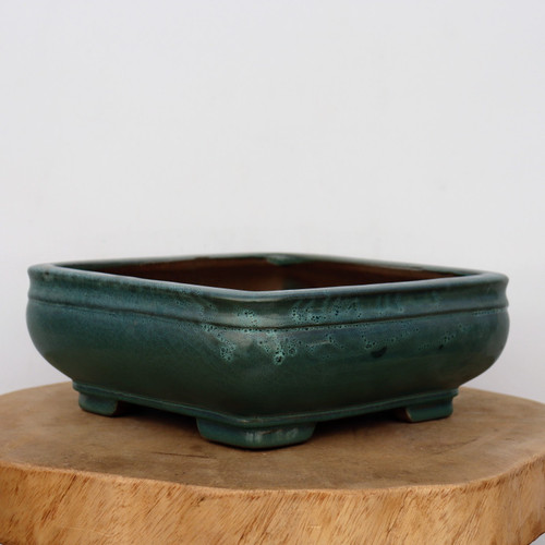 An artisanal nine-inch Yixing bonsai planter is featured in a close-up, showcasing its rich teal-green glaze. The rectangular ceramic pot has softly rounded corners, slightly bowed sides, and a prominent top rim. Its glaze is deep and lustrous, with subtle variations and light, speckled "frosted" textures near the upper edges that highlight its handmade character. 

The planter is elevated by four sturdy, blocky feet at each corner. It is positioned on a rustic, light-colored wooden slab with visible natural grain and bark texture along the edge. The entire scene is set against a clean, minimalist off-white background, emphasizing the elegant form and vibrant color of the glazed bonsai pot.