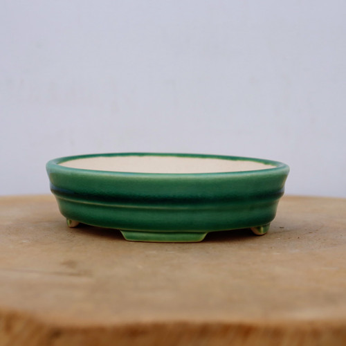 A shallow, oval-shaped Yixing glazed bonsai pot rests on a light-colored wooden surface against a plain white background. The ceramic planter features a vibrant, multi-toned emerald green glaze with darker green detailing along its stepped, double-rimmed exterior. The interior of the pot is a contrasting pale cream color. It is supported by small, integrated feet that lift it slightly off the surface. This classic green glazed bonsai pot is designed for small to medium-sized bonsai trees, blending traditional craftsmanship with a clean, professional aesthetic.