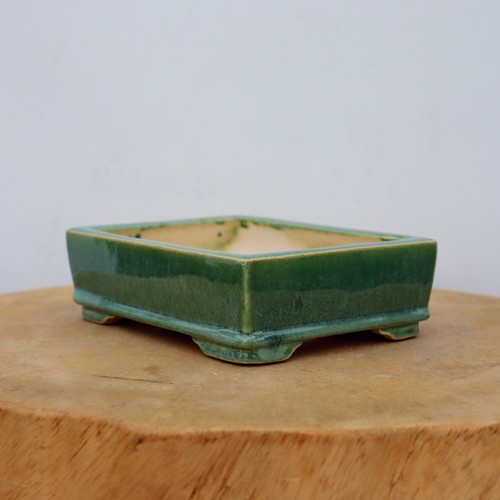 A rectangular Yixing glazed bonsai pot in a deep forest green color is showcased in a close-up, eye-level shot. The ceramic planter features a glossy finish that catches the light, with a slightly flared rim and a matching flared base. It rests on four short, rectangular feet at each corner, providing a stable foundation and allowing for drainage.

The interior of the pot reveals a pale, off-white unglazed ceramic surface. This high-quality Yixing planter is displayed on a thick, rustic wooden block with a visible natural grain, set against a plain, neutral-toned wall. This simple setting emphasizes the pot's elegant form and rich, jewel-toned color. The image highlights the pot's handcrafted quality and its suitability for a variety of bonsai trees, blending traditional Chinese pottery techniques with a vibrant, modern aesthetic.