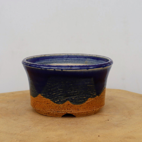 A side-view photograph of a round, American handmade ceramic bonsai pot by artist Sean Guo. The pot features a striking two-tone finish: the upper half and rim are coated in a rich, glossy cobalt blue glaze that flows down in a jagged, mountain-like silhouette over the base. The lower half of the pot exposes a textured clay body in a warm, earthy ochre-orange hue, marked by subtle horizontal throwing rings and fine dark speckles. This artisanal bonsai container has a slightly flared rim and small arched drainage cutouts at the foot. It sits centered on a light-colored wooden surface against a neutral, off-white background, highlighting the rugged yet refined craftsmanship of the piece.