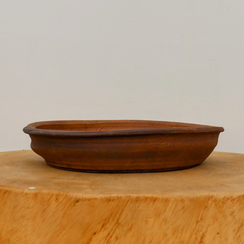A shallow, oval-shaped unglazed bonsai pot handcrafted by American artist Paul Olson sits centered on a light-colored, natural wood pedestal. The pot is made from a rustic, earthy terracotta clay with a rich, reddish-brown color and subtle darker, scorched-looking accents around the flared rim. Its design features a low profile with a slight horizontal ridge running around the middle of its textured exterior, highlighting its artisanal quality. The pot is photographed from a side-view against a clean, off-white background, emphasizing the natural tones and handcrafted details of the ceramic piece.