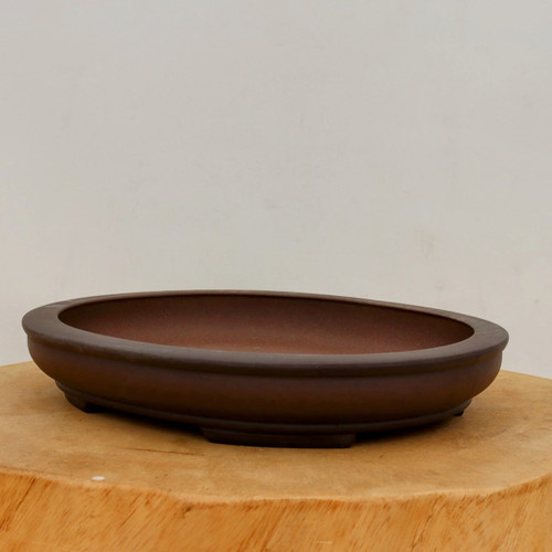 An elegant, high-quality Yixing unglazed bonsai pot (model 2451) featuring a deep, dark brown matte finish. The pot is shaped in a classic shallow oval with a wide, rounded rim that slopes slightly inward. It rests on small, integrated feet designed to provide essential drainage and air circulation for a bonsai tree. The pot is displayed on a rustic, light-toned wooden pedestal, creating a striking contrast that emphasizes its smooth texture and traditional craftsmanship. The background is a clean, neutral off-white, making this professional-grade ceramic piece the central focus.