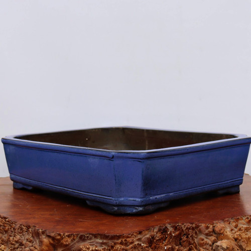 A detailed view of a Yixing 2285 glazed bonsai pot, characterized by its deep cobalt blue color and elegant rectangular shape with indented, canted corners. The ceramic pot features a smooth finish with subtle mottling and two parallel horizontal decorative ridges encircling the upper and lower edges. It sits on short, integrated feet, resting atop a polished, reddish-brown wood slab with a highly textured, natural bark edge. This handcrafted container is set against a neutral, off-white background, highlighting the rich blue glaze and traditional craftsmanship characteristic of Yixing pottery, making it a sophisticated choice for various bonsai tree styles.