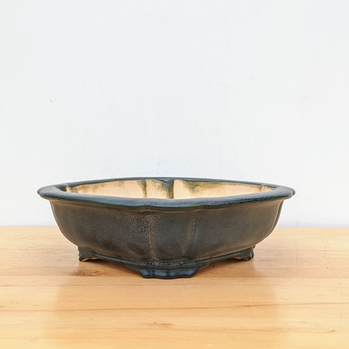 A Yixing No. 2323H glazed ceramic bonsai pot, featuring a deep, matte forest green finish with a subtle mottled texture. The pot has an elegant, scalloped rectangular shape with soft, rounded corners and a flared rim. It is elevated on four small, decorative feet that mirror the scalloped design of the body. The interior of the pot is unglazed, showing the natural light tan color of the clay. The pot is centered on a light-colored wooden surface against a clean, plain white background, highlighting its form and earthy color palette.