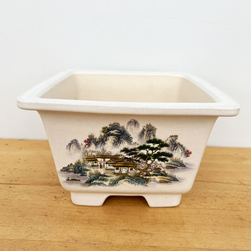 A high-angle, front-facing shot shows a 9-inch glazed Yixing bonsai pot, ideal for cascade or semi-cascade bonsai styles. The pot is a creamy off-white color with a square shape, a wide flared rim, and four small rectangular feet at the base. It sits on a light-grained wooden tabletop against a plain white background. 

The front side of the pot features an intricate, hand-painted traditional Chinese landscape scene. The painting depicts mist-covered, rugged mountains in the background with a small village of thatched-roof houses nestled in the valley. In the foreground, a large, gnarled pine tree stands next to smaller trees with pink blossoms, reminiscent of cherry or plum blossoms. The scene is rendered in a "Shan Shui" style with a palette of earthy greens, blues, browns, and subtle pops of red and pink, capturing a peaceful, ancient wilderness aesthetic.