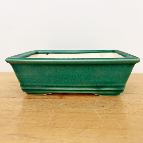 A detailed side-on view of a 7-inch glazed rectangle Yixing ceramic bonsai pot from Bonsai Outlet. The pot is finished in a vibrant emerald green glaze with a delicate crackle texture, also known as crazing, which adds a vintage aesthetic. Its design features a slightly flared rim and a decorative double-beaded indentation along the bottom edge, resting on four small, integrated feet that elevate the pot for proper drainage. The interior of the pot shows a contrasting light cream-colored ceramic. It is displayed on a light brown wooden tabletop against a plain white background, highlighting its clean lines and rich color, making it an ideal choice for housing a bonsai tree.