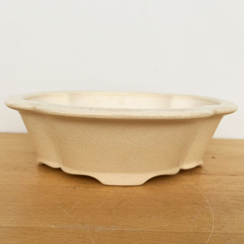 A 10-inch cream-colored glazed Yixing ceramic bonsai pot sits centered on a light-toned wooden surface against a minimalist white background. The pot features a distinctively elegant scalloped or lobed oval shape with a gently flared rim and a series of shallow, integrated wavy feet at the base for elevation. The cream glaze is characterized by a delicate crackle or crazing pattern across the entire exterior, providing a sophisticated, antique aesthetic. The low-profile design is captured from a side-angle view, highlighting the pot's graceful curves and fine craftsmanship, ideal for displaying a mature bonsai tree.