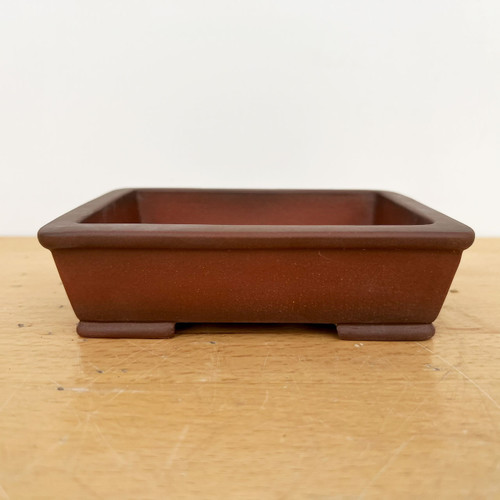 A side-profile view of a 5.5-inch rectangular Yixing ceramic bonsai pot. The pot is unglazed, showcasing a smooth, matte texture in a deep reddish-brown, earthy clay tone. Its design features a wide, flat rim and sides that slope gently inward toward the base. The pot is elevated by four small, integrated rectangular feet at the corners, providing drainage space underneath. It is positioned centrally on a light-colored wooden surface with a visible natural grain, set against a minimalist, solid off-white background.