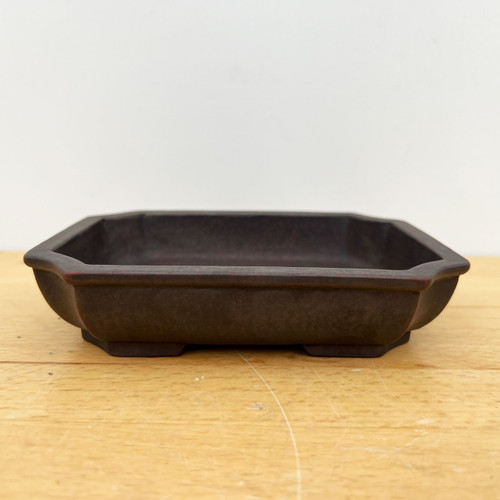An eye-level, front-facing photograph captures a 6-inch unglazed rectangular Yixing ceramic bonsai pot. The pot is a deep, rich chocolate brown color with a matte, earthy texture characteristic of high-quality Yixing clay. It features an elegant elongated octagonal shape with softly indented corners and a clean, flat top rim that has a subtle outer lip. The shallow container sits on four small, integrated feet, providing a slight elevation. Positioned centrally on a light-toned, natural wood grain surface against a plain, off-white background, the image highlights the pot's minimalist and traditional design, making it an ideal choice for various bonsai tree styles that require a breathable, unglazed ceramic home.