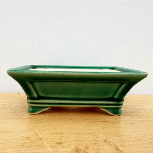 A front-facing, eye-level photograph of a small, rectangular forest green glazed Yixing ceramic bonsai pot. The pot features a glossy, deep jade green glaze that pools in the horizontal molding lines and around the base, creating rich, darker emerald tones that contrast with the lighter green on the flared upper rims. The container has a traditional architectural design with four integrated feet that lift it slightly off the surface. 

Inside the pot, a light beige, unglazed ceramic finish is visible. The bonsai pot sits centrally on a light-toned wooden tabletop with visible grain and fine scratches, set against a plain, off-white wall. This classic ceramic container is designed for small bonsai trees or succulents, showcasing the craftsmanship of Yixing pottery and offering a durable, aesthetic home for miniature plants.