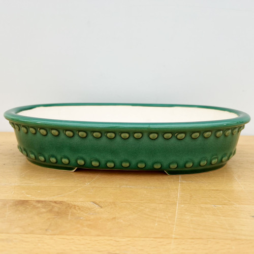 A detailed, side-profile view of a 12-inch drum-style oval Yixing ceramic bonsai pot. The pot is finished in a rich, vibrant forest green glaze that features an intricate, fine-line crackle texture. A distinctive design element is the two parallel rows of raised, circular ceramic studs or rivets circling the exterior body, giving it a classic drum-like appearance. 

The pot has a shallow, oval shape with a smooth, rounded upper rim and sits on small, integrated feet that subtly elevate it. The interior is unglazed, revealing a clean, pale cream-colored clay body. The pot is shown resting on a light-toned wooden tabletop against a plain, neutral white background, highlighting its color and craftsmanship.