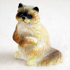 A detailed miniature Ragdoll cat figurine designed as a decorative bonsai accessory or fairy garden ornament. The small statue depicts a fluffy Ragdoll cat sitting upright on its haunches with its front paws tucked neatly against its chest in an alert, curious pose. It features characteristic pointed markings, including a dark seal-brown face mask, black-tipped ears, and a thick, bushy brown tail that contrasts with its creamy white and light tan long-haired coat. The figurine is accented with bright, vivid blue eyes and a textured surface that realistically mimics the look of tufted fur. It is shown against a plain white background, highlighting its intricate details for use in miniature landscaping.