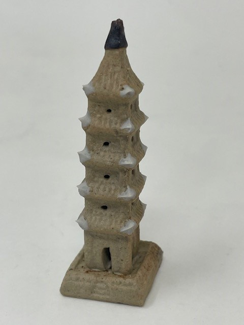 A detailed miniature Chinese pagoda figurine, styled as a traditional "mudman" ancient tower intended for bonsai decoration. The ceramic structure features five distinct tiers, each with flared eaves accented by pointed, off-white tips at the corners. Crafted from a textured, earthy tan clay, the tower includes small, dark circular indentations representing windows on each level. At the very bottom, a small arched entryway is carved into the first floor above a solid, square-shaped pedestal base with faint geometric relief patterns. The pagoda is capped with a dark brown, conical finial at the peak. The figurine is centered against a plain, off-white background, highlighting its handcrafted details and traditional aesthetic.