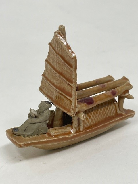 A high-resolution, close-up photograph of a miniature Chinese mudman figurine depicting a traditional skiff boat, designed as a decorative accessory for bonsai trees. The handcrafted ceramic boat is finished in a glossy, light brown or tan glaze. It features a tall, vertical sail with horizontal ribs, textured to resemble woven bamboo or fabric. 

At the bow of the skiff sits a small, unglazed grey mudman figure, hunched forward as if rowing or tending to the vessel. The middle section of the boat carries several thick, horizontal log-like structures, some of which are accented with patches of deep reddish-purple glaze. A section of the boat's side features a cross-hatched, lattice-like pattern. The entire figurine is set against a clean, neutral white background, emphasizing the intricate textures and traditional craftsmanship of the piece.