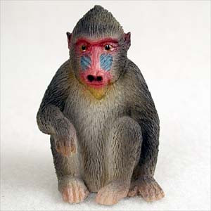 A detailed miniature figurine of a mandrill, designed as a decorative accessory for a bonsai tree or terrarium. The monkey is depicted in a seated, upright pose with its hands resting near its feet. It features realistic textures and colors, including a dark grayish-brown coat of fur, a pale underbelly, and the species' characteristic vibrant face with a bright red nose and muzzle, blue ridged cheeks, and a small yellowish beard. The figurine is shown against a plain white background, highlighting its intricate sculpted details.