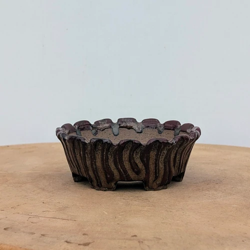A side-view studio photograph of a unique, handmade ceramic bonsai pot by artist Brandon Besserer (No. 9). The shallow, round pot is crafted from a coarse, light-brown stoneware and features a decorative scalloped rim. The exterior is characterized by deep, vertical wavy ridges. A dark, glossy reddish-brown glaze has been applied in a vertical, striped pattern over the textured surface, leaving parts of the matte, tan clay exposed for a rustic, organic look. The pot rests on four small, subtle feet on a flat wooden surface against a minimalist white backdrop, highlighting its artisanal craftsmanship and earthy aesthetic.