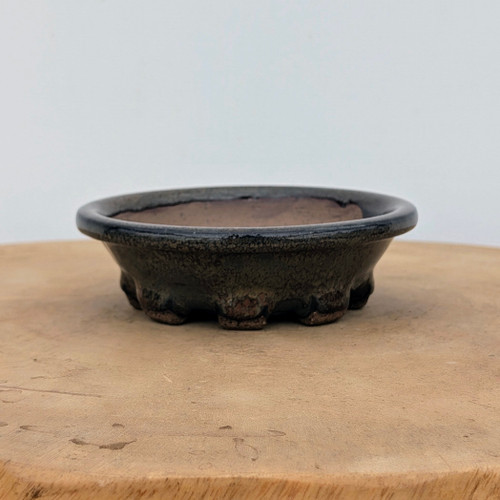 A detailed side-view photograph of a handmade ceramic bonsai pot, identified as Brandon Besserer No. 10. This round, shallow pot features a wide, flared rim and sits on several small, chunky feet that provide elevation. The exterior is coated in a dark, metallic charcoal glaze with a subtle, mottled texture, while the interior and the underside of the feet showcase the unglazed, reddish-brown natural clay. The pot is positioned in the center of a light-colored wooden display stand against a neutral, off-white background, highlighting its artisanal craftsmanship and earthy aesthetic.