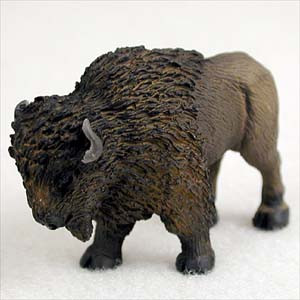 A highly detailed miniature figurine of an American bison, commonly referred to as a buffalo, designed as a decorative bonsai tree accessory. The figurine features a realistic, hand-painted texture, with thick, shaggy dark brown fur covering the massive head, shoulders, and hump, transitioning to a smoother, lighter tan-brown finish on the rear half. The bison is depicted in a natural, sturdy stance with its head slightly lowered, showcasing a small, curved grey horn and dark, detailed hooves. It is photographed against a clean, white background to highlight the intricate sculpting and lifelike appearance of the small-scale animal.