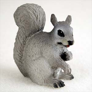 A detailed miniature figurine of a grey squirrel, designed as a decorative bonsai tree accessory. The squirrel is depicted sitting upright, facing right, with its small front paws held near its mouth as if nibbling. Its large, bushy tail is elegantly curled upwards behind its back. The figurine showcases a realistic fur-like texture with black-painted eyes, nose, and paws, and a hint of a pink tongue. It is shown against a neutral, off-white background.