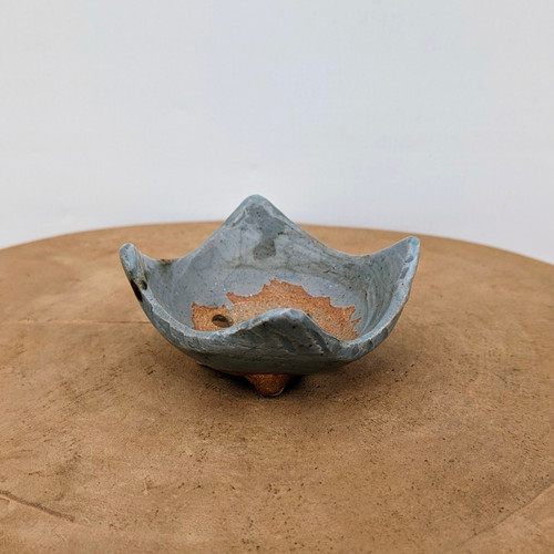 An American handmade ceramic bonsai pot by artist Bruce Lenore sits centered on a light brown wooden surface. The small pot has an organic, star-like shape with an irregular, undulating rim that rises into four soft points. A cool, mottled bluish-gray glaze covers the exterior and much of the interior, contrasting with the warm, unglazed reddish-brown clay visible at the very center of the bowl and on the three small, rounded feet underneath. A single drainage hole is clearly visible in the bottom of the pot. The background is a clean, neutral white, emphasizing the pot's textured surface and unique artisan design.