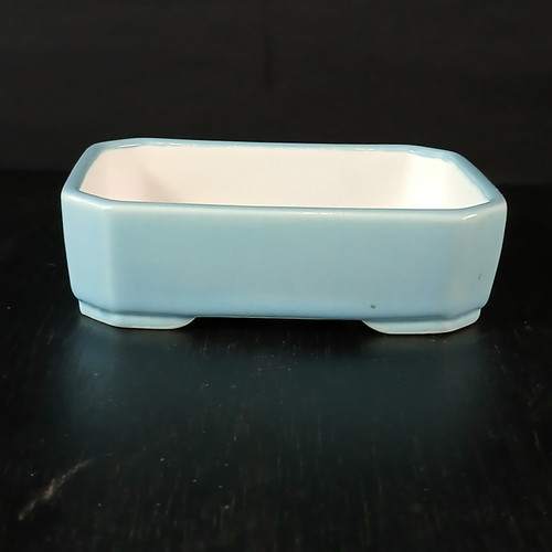 A 5-inch rectangular Yixing ceramic bonsai or succulent pot with a vibrant light blue glazed exterior. The pot's design includes softly clipped corners and a slight outward flair towards the top rim, providing a classic silhouette. The interior is finished in a clean white glaze, offering a stark contrast to the colorful outer surface. Positioned on a dark, polished wood-like surface against a deep black background, the pot's glossy finish and elegant form are accentuated. This miniature planter is designed with small drainage-ready feet at the base, making it a functional and aesthetic choice for housing a small bonsai tree or a selection of succulents.