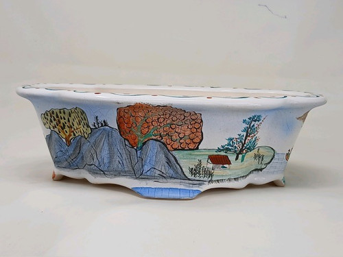This 10-inch rectangular Yixing bonsai pot features a white glazed ceramic finish with a delicate crazing pattern throughout. The pot has a flared, scalloped rim and is supported by four small feet at the corners. A hand-painted landscape in a traditional style wraps around the front and sides. On the left, craggy, stylized mountains in shades of blue and gray with black outlines rise upward, topped with a bright yellow and green tree. The center of the scene showcases a large tree with textured, orange foliage and a smaller white house with a slanted red roof nestled on a green landscape. To the right, additional trees with blue and green leaves are painted against a pale background. The scalloped rim is delicately decorated with a series of small red dots and blue line patterns, and a central base element is accented with a blue cross-hatched design. This artistic bonsai pot, identified as model No. 2127H, is a fine example of a decorative, hand-painted container designed for displaying a bonsai tree.